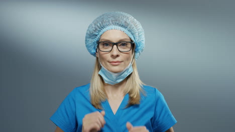 Portrait-shot-of-the-young-attractive-blond-Caucasian-female-physician-in-the-blue-hat-and-glasses-taking-off-her-mask-from-the-mouth-and-smiling-happily.-Close-up.